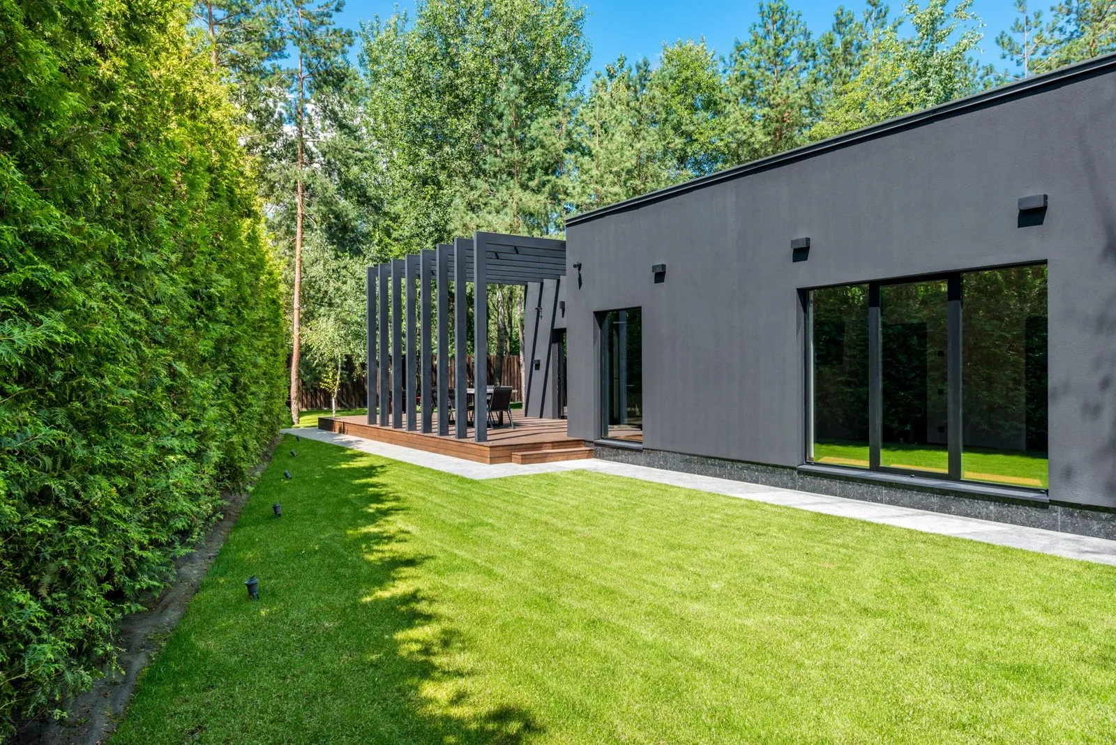 Low dark rendered house with a pergola and deck at the end of a long lawn, tall conifer hedge alongside.