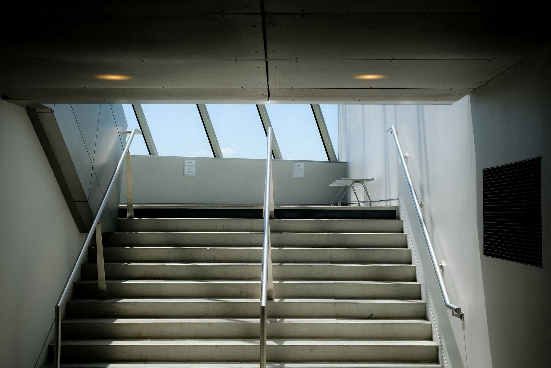 Wide concrete stair with steel handrails rising towards a band of sloping roof glazing.